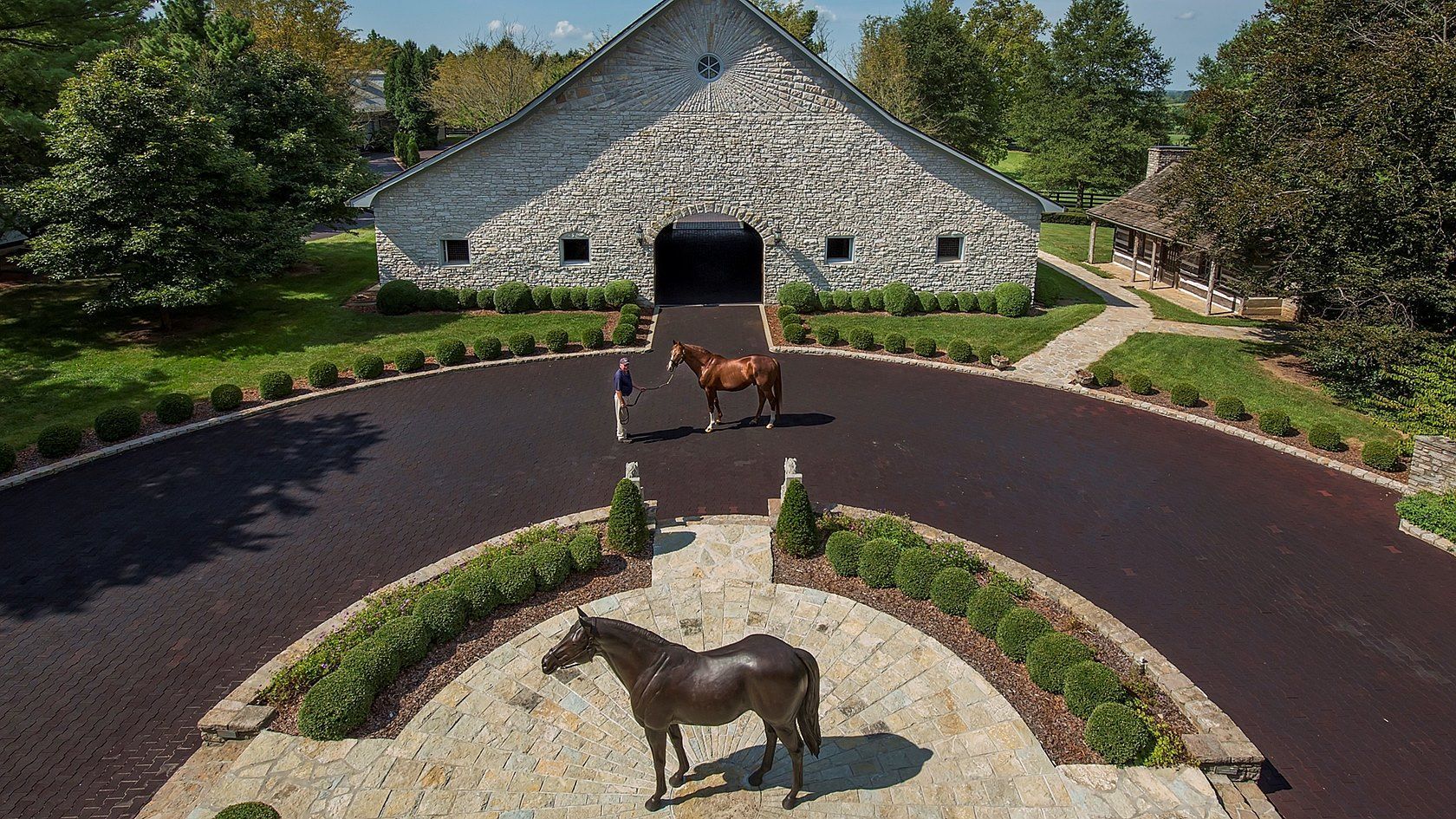 Limestone horse barn with a chestnut horse on the circular drive and a bronze horse sculpture in the foreground